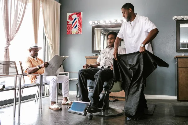 Barbeiro coloca a capa em um cliente na cadeira enquanto outro cliente lê jornal na área de espera de uma barbearia