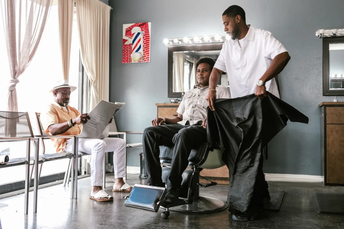 Barbeiro coloca a capa em um cliente na cadeira enquanto outro cliente lê jornal na área de espera de uma barbearia