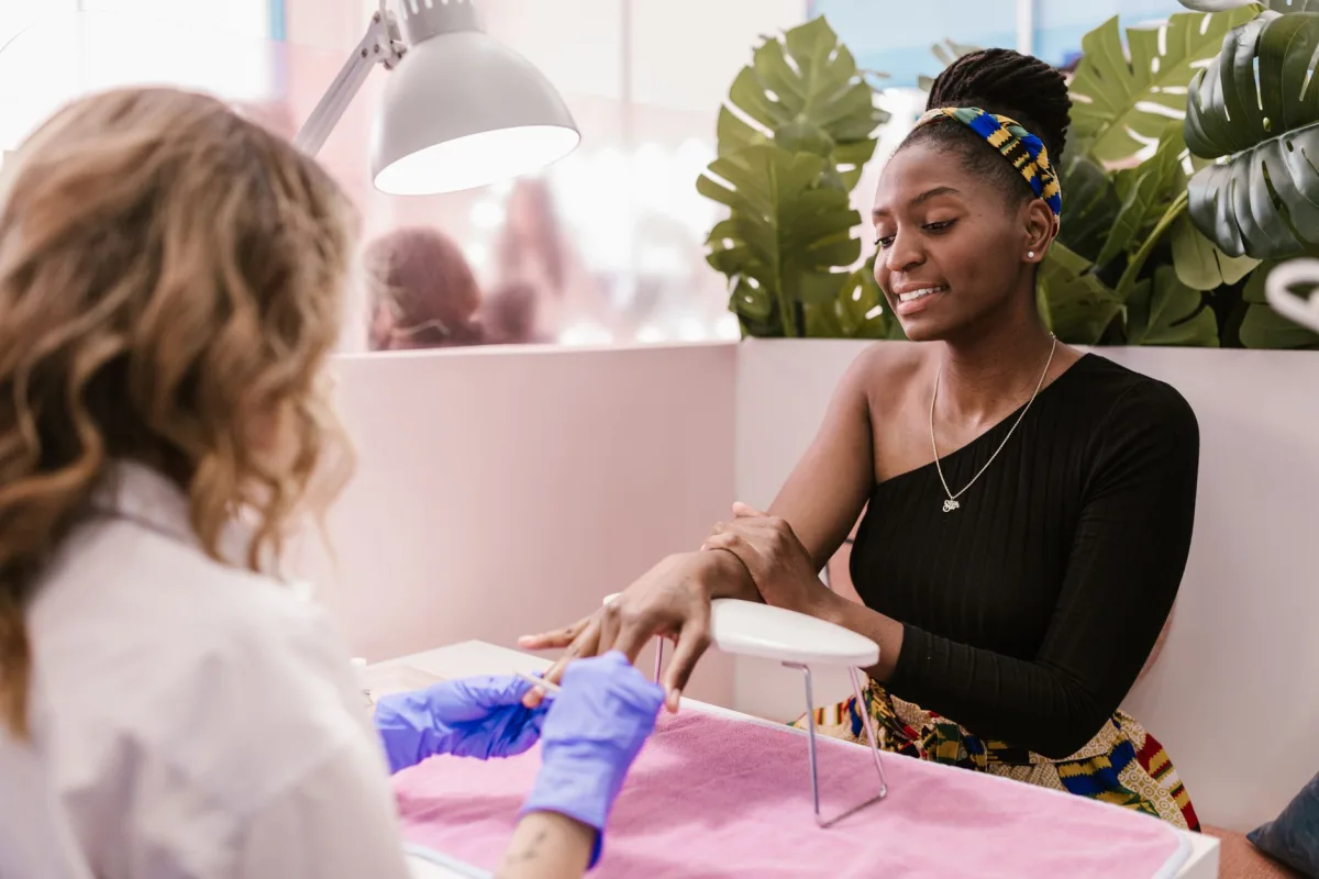 Manicure de luvas roxas atende uma cliente negra sorridente em uma bancada de esmalteria