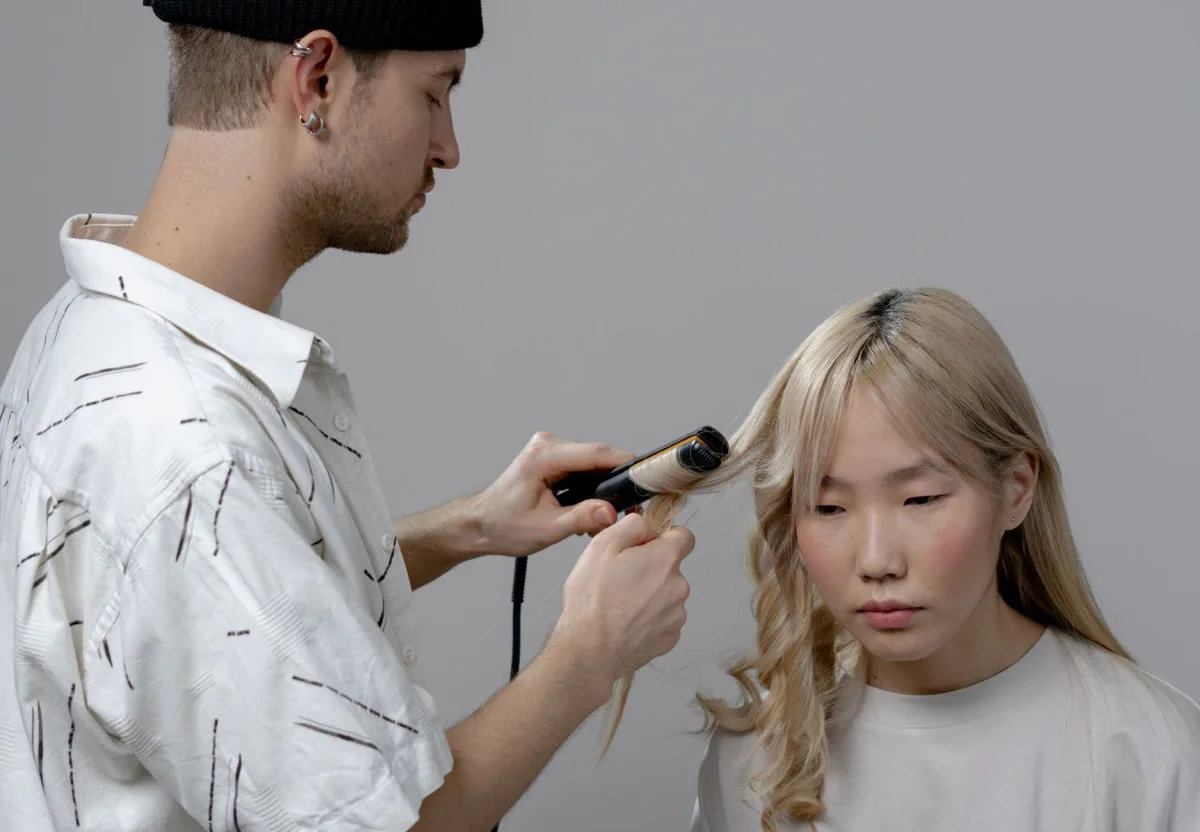 Cabeleireiro jovem de touca e camisa estampada modela com babyliss o cabelo loiro platinado de uma cliente asiática