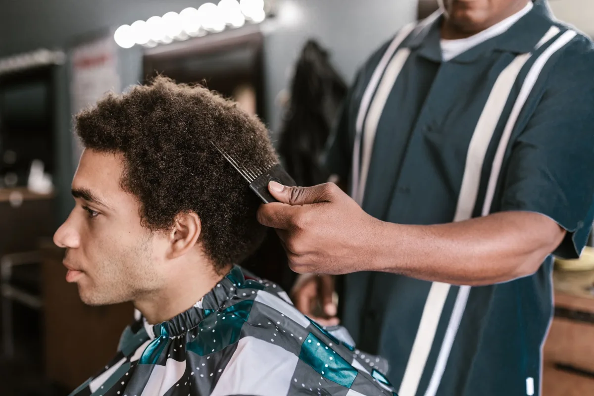 Barbeiro negro penteia com pente garfo o cabelo crespo de um cliente jovem sentado na cadeira de uma barbearia