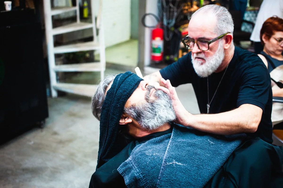 Barbeiro grisalho de óculos e barba branca apara a barba de um cliente de cabelo grisalho reclinado na cadeira de uma barbearia