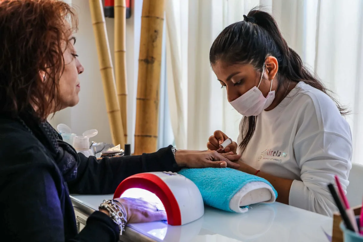 Manicure jovem de cabelo preso e máscara faz as unhas de uma cliente madura de cabelo ruivo, com cabine de LED sobre a bancada do salão