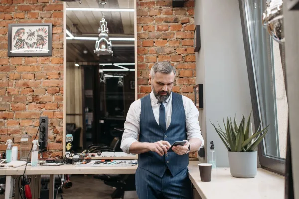 Dono de barbearia de colete e gravata consulta o celular em pé no salão, com parede de tijolos ao fundo